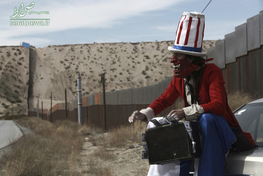 یک معترض با لباس عمو سام و چمدانی پر از پول در مقابل دیوار مرزی آمریکا و مکزیک